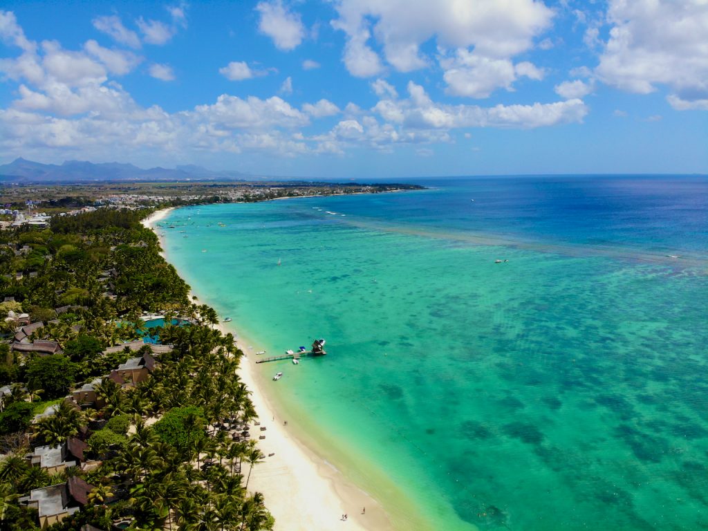 Auf dem Bild sieht man die Lagune von Trou aux Biches auf Mauritius aus der Vogelperspektive. Eine Drohnenaufnahme. Man sieht die Lagune mit dem türkisen Wasser, den Strand und ein Stück vom Beachcomber Hotel links daneben. Im Hintergrund sieht man eine Bergkette und eine Landzunge mit einer Dornenkante. 

Der Strand von Trou aux Biches aus der Vogelperspektive. Foto: Sascha Tegtmeyer