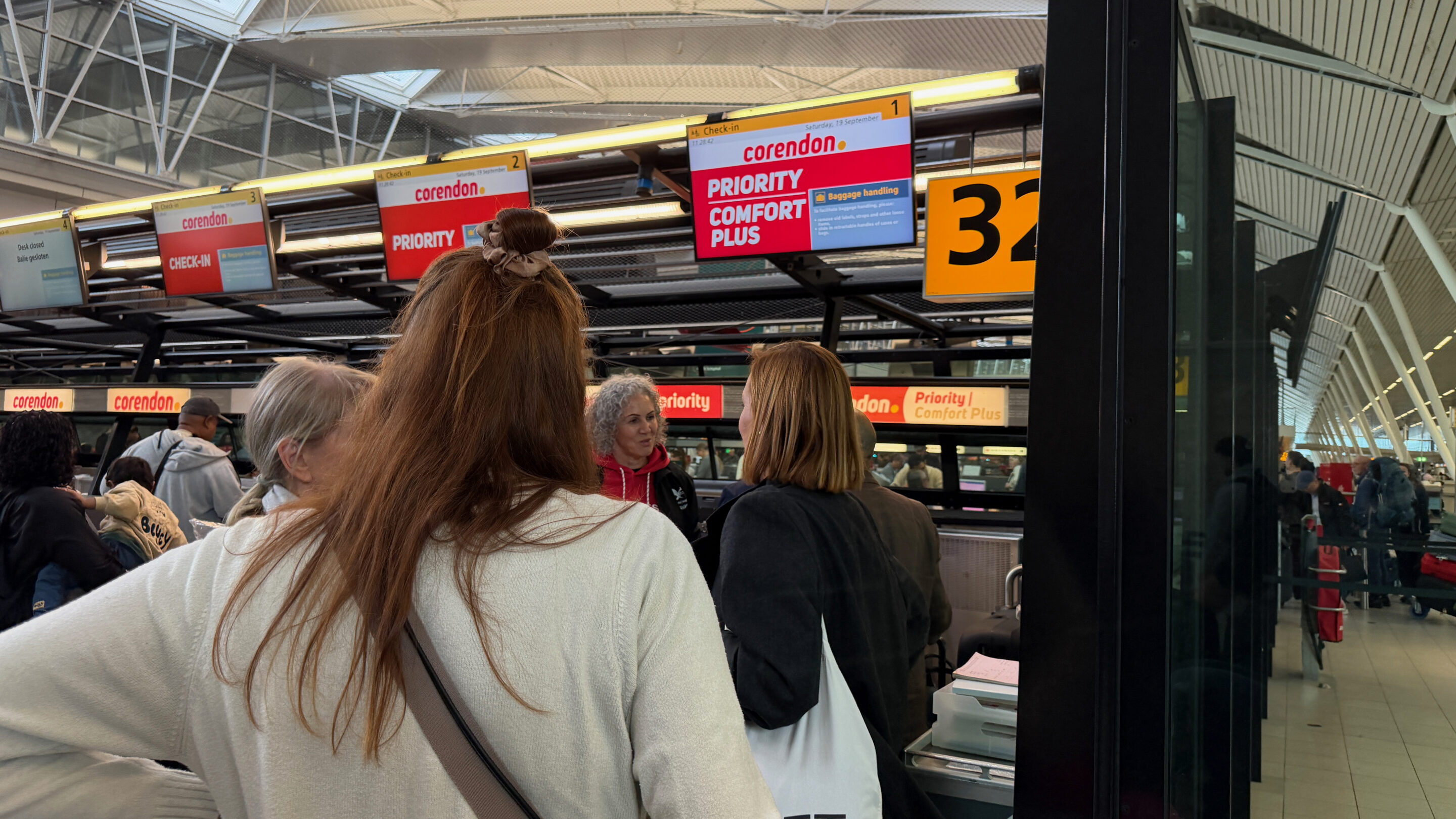 Flughafen Amsterdam Schiphol – meine Erfahrungen mit gut sechs Stunden Aufenthalt 14 Reisende von hinten vor einem Corendon-Priority-Check-in-Schild in Schiphol