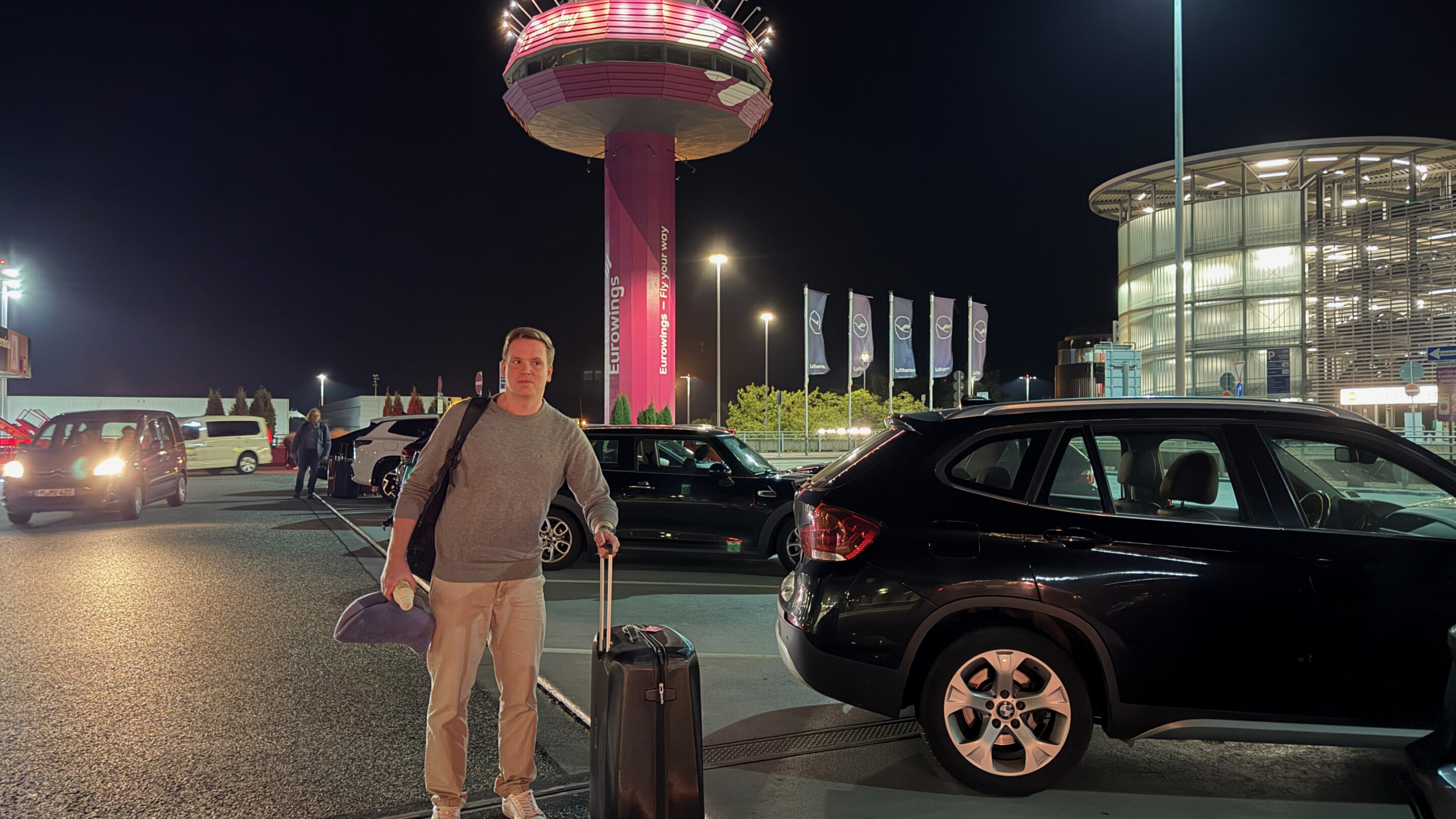 Sascha mit Koffer vor dem Flughafen Hannover und pink beleuchtetem Turm